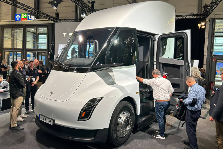 A imagem mostra um caminhão elétrico de cor branca em uma exposição. O veículo possui um design moderno, com uma cabine ampla e janelas grandes. Há várias pessoas ao redor, algumas interagindo com o caminhão, enquanto outras observam. O ambiente parece ser um espaço de evento, com iluminação adequada e estruturas metálicas ao fundo.