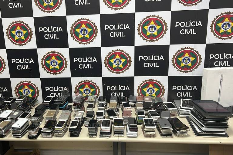 A imagem mostra uma mesa repleta de celulares e dispositivos móveis, dispostos em várias fileiras. Ao fundo, há um painel com o logotipo da Polícia Civil, composto por quadrados pretos e brancos, com emblemas da polícia. A cena sugere uma operação policial relacionada a apreensões de eletrônicos.