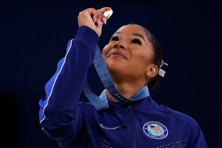 Uma atleta está olhando para cima, segurando uma medalha em sua mão. Ela usa um agasalho azul com o emblema da equipe olímpica dos Estados Unidos. A atleta parece emocionada e está com um sorriso no rosto, enquanto a medalha brilha sob a luz.
