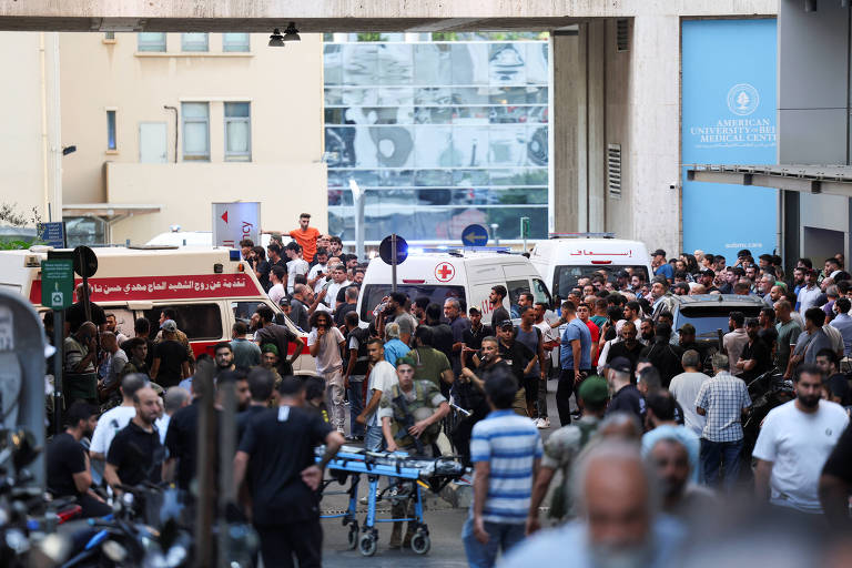 A imagem mostra uma grande multidão reunida em uma área urbana, com várias ambulâncias visíveis ao fundo. As pessoas parecem estar se aglomerando em torno das ambulâncias, e há sinais de atividade intensa. Algumas pessoas estão usando roupas casuais, enquanto outras parecem estar em uniformes. O ambiente é urbano, com edifícios ao fundo e uma atmosfera de urgência.