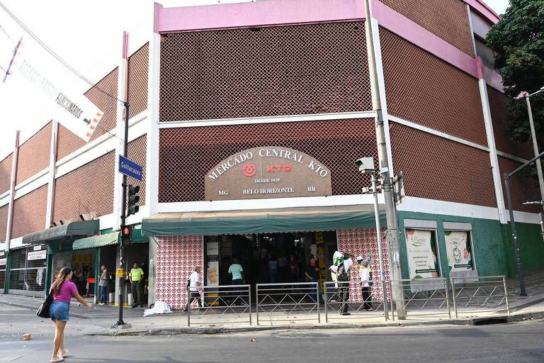 A imagem mostra a fachada do Mercado Central KTO, localizado em Belo Horizonte, Brasil. O edifício tem uma estrutura de tijolos expostos e detalhes em rosa. Na entrada, há uma faixa com o nome do mercado e informações sobre sua fundação em 1929. Algumas pessoas estão na calçada em frente ao mercado, e há árvores e postes de iluminação ao redor.