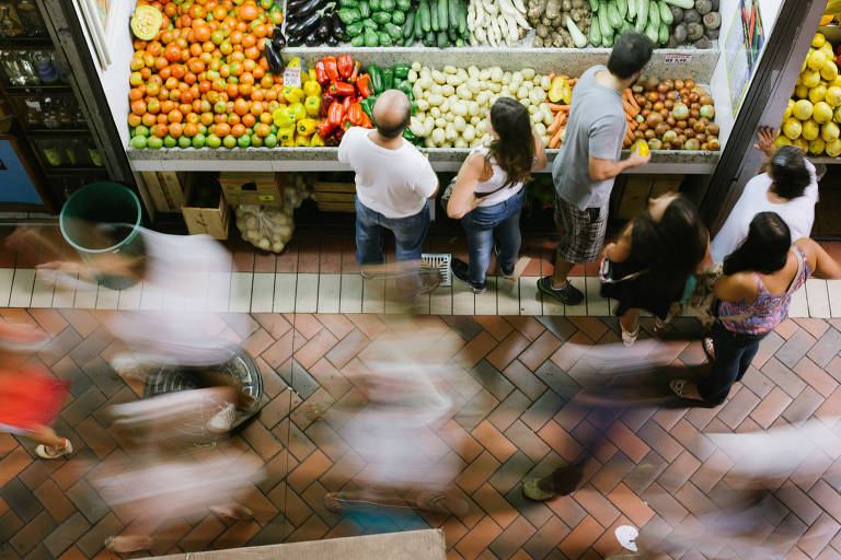 A imagem mostra uma feira de frutas e verduras, com uma variedade de produtos frescos dispostos em uma bancada. Há pessoas em pé, observando as mercadorias, enquanto outras estão em movimento ao redor. As frutas incluem laranjas, pimentões, abobrinhas e batatas. O chão é de ladrilhos em padrão de espinha de peixe, e há uma lixeira verde ao fundo.