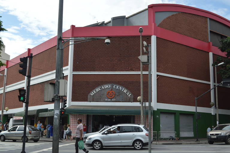 A imagem mostra a fachada do Mercado Central, um edifício de dois andares com uma estrutura de tijolos vermelhos e detalhes em branco e rosa. O letreiro 'MERCADO CENTRAL' está visível na entrada, junto com a data 'DESDE 1929'. Há semáforos visíveis, um com luz verde, e várias pessoas e carros na rua em frente ao mercado.