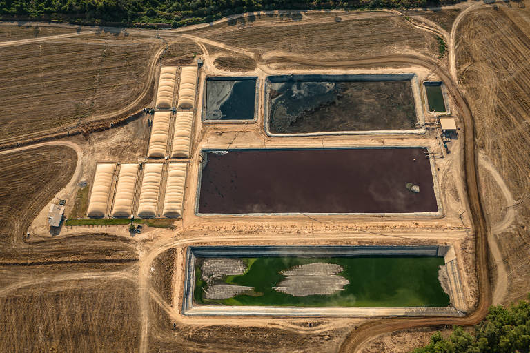 Imagem aérea de um complexo de tratamento de água, com várias lagoas de diferentes cores, incluindo uma lagoa verde, uma lagoa marrom e uma lagoa escura. Há também estruturas cobertas em forma de arcos ao lado das lagoas. O terreno ao redor é predominantemente seco e árido, com algumas áreas de vegetação ao fundo.