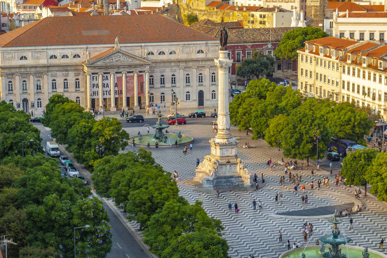 A imagem mostra uma praça ampla com um monumento central elevado, cercado por árvores verdes. Ao fundo, há um edifício histórico com uma fachada clássica. A praça é pavimentada com um padrão de ondas e está cheia de pessoas caminhando. Vários carros estão estacionados nas proximidades, e o céu está claro, indicando um dia ensolarado.