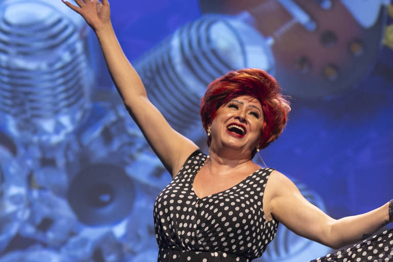 Uma mulher com cabelo vermelho e vestido de bolinhas pretas e brancas está se apresentando em um palco. Ela está sorrindo e levantando os braços, enquanto uma imagem de fundo mostra microfones e instrumentos musicais. A cena transmite uma atmosfera vibrante e alegre.