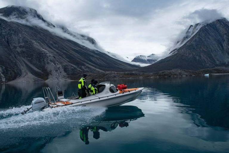 A imagem mostra um barco motor navegando em um lago cercado por montanhas. O céu está nublado e há neblina nas montanhas. No barco, há três pessoas vestindo coletes amarelos. O reflexo das montanhas e do barco pode ser visto na água calma do lago.