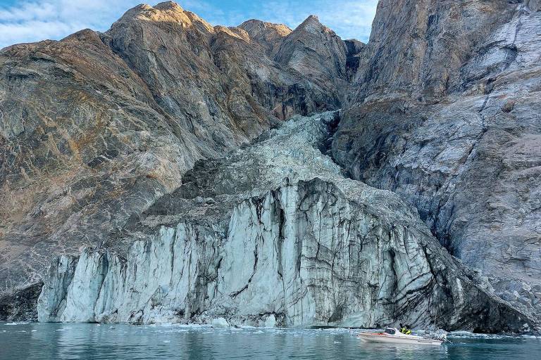 A imagem mostra um grande glaciar em uma montanha, com rochas expostas ao fundo. A superfície do glaciar é de cor azulada e há uma pequena embarcação na água em frente ao glaciar. O céu está parcialmente nublado, e as montanhas ao fundo são íngremes e rochosas.