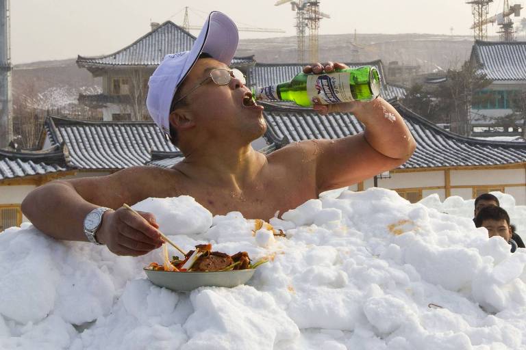 Homem sem camisa, enterrado num monte de neve, bebe cerveja