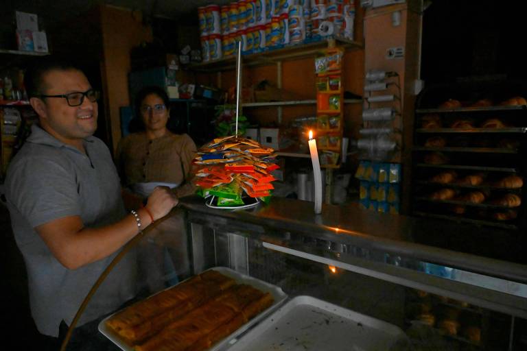 A imagem mostra um homem sorridente em uma loja durante um apagão, segurando um suporte com pacotes de salgadinhos empilhados. Ao fundo, uma mulher observa. Uma vela acesa ilumina a cena, enquanto prateleiras com produtos e pães são visíveis ao fundo.
