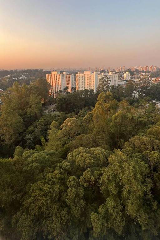 A imagem mostra uma vista panorâmica de uma área urbana ao entardecer. No primeiro plano, há uma densa vegetação verde, com árvores altas. Ao fundo, destacam-se prédios residenciais de cor clara, com janelas refletindo a luz do sol. A cidade se estende ao longe, com mais edifícios visíveis sob um céu que gradualmente muda de cor, do azul para tons de laranja e rosa, indicando o pôr do sol.