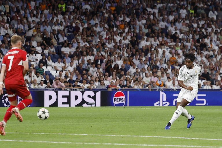 A imagem mostra um jogador do Real Madrid, vestido com um uniforme branco, chutando a bola em direção ao gol. Um jogador adversário, com um uniforme vermelho e o número 7 nas costas, está se afastando. Ao fundo, há uma grande quantidade de torcedores nas arquibancadas, predominantemente vestindo roupas claras. Um painel publicitário da Pepsi é visível na parte inferior da imagem.
