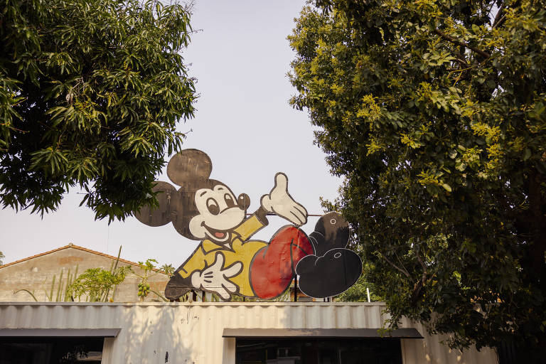 A imagem mostra um mural de Mickey Mouse, um personagem icônico da Disney, em uma parede. Mickey está deitado, com um sorriso, vestindo uma camisa amarela e calças vermelhas. O fundo é composto por árvores verdes e um céu claro. A parte inferior da imagem mostra uma estrutura que parece ser um edifício.