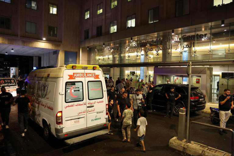 A imagem mostra uma ambulância estacionada em frente a um hospital à noite. Há um grupo de pessoas ao redor da ambulância e na entrada do hospital, com algumas pessoas se movendo. O ambiente é iluminado por luzes artificiais, e o prédio do hospital possui grandes janelas.