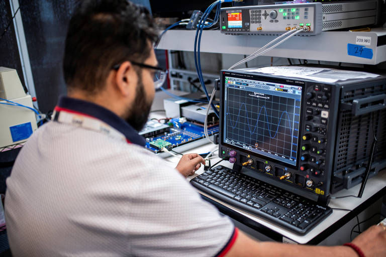 Um homem está sentado em frente a um computador em um laboratório de eletrônica, operando um osciloscópio. O osciloscópio exibe um gráfico de ondas senoidais. O ambiente é equipado com diversos dispositivos eletrônicos e cabos. O homem usa óculos e uma camisa com detalhes em vermelho e azul.
