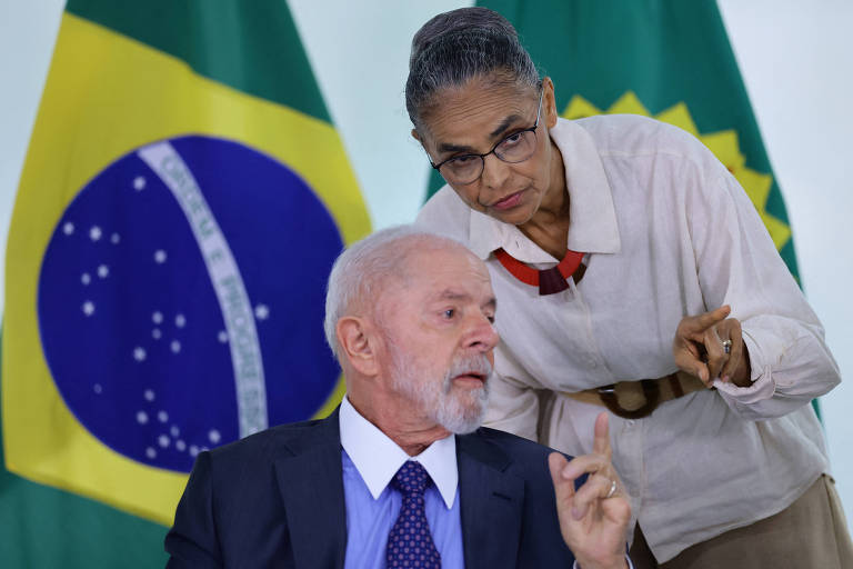 Lula, de terno azul escuro, gravata azul, camisa azul clara com colarinho branco, está sentado e gesticula e conversa com Marina Silva, que está de pé. Ela usa camisa de linho branca com cinto e colar indígena e saia bege. Está de pé se curva para falarcom o presidente.