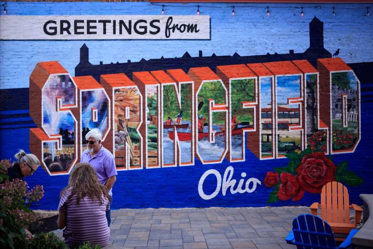 A imagem mostra um mural colorido com a frase 'Greetings from Springfield' em letras grandes e estilizadas, com a palavra 'Springfield' destacada em laranja. Abaixo, está escrito 'Ohio' em letras brancas. O mural apresenta várias imagens representativas da cidade, e há pessoas em primeiro plano, uma delas segurando um celular. O fundo é azul.