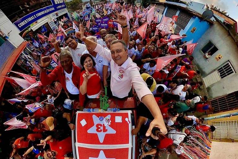 A imagem mostra um comício animado com uma grande multidão reunida em torno de um carro. As pessoas estão segurando bandeiras e cartazes, muitos vestindo roupas vermelhas. No centro, Rui Costa sorri e faz gestos de vitória, enquanto o sol se põe ao fundo. O carro tem o número 13 visível em seu teto, e a cena é cercada por casas e bandeirinhas coloridas