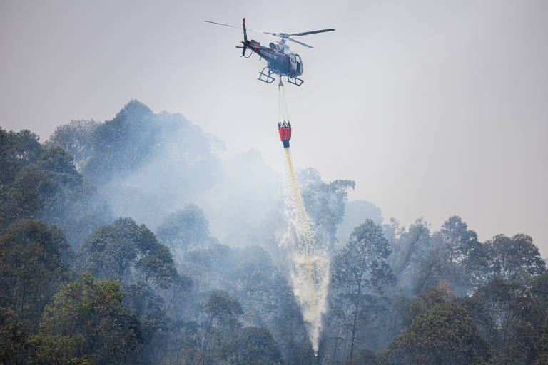 Um helicóptero está sobrevoando uma área florestal, despejando água para combater um incêndio. A cena é envolta em fumaça, com árvores visíveis na parte inferior da imagem. O helicóptero está equipado com um balde suspenso, do qual a água está sendo liberada.