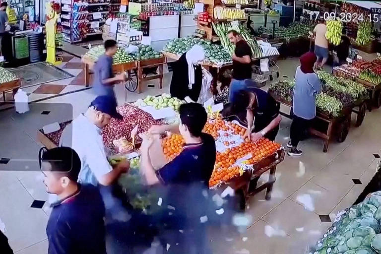 A imagem mostra o interior de um mercado, onde várias pessoas estão interagindo entre si. Há mesas com frutas e vegetais dispostos em várias cores. No centro, é possível ver um homem com pedaços de roupa explodindo ao seu redor.