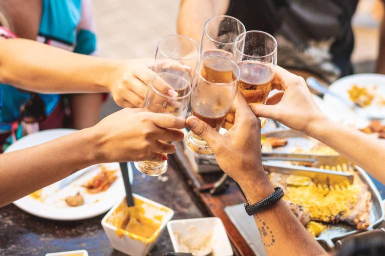 Mãos segurando copos de chopp e brindando, com uma mesa cheia de restos de comida ao fundo
