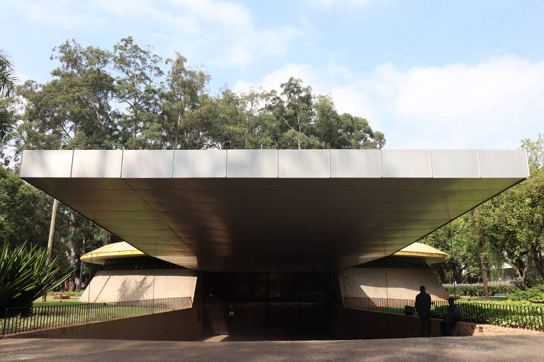 A imagem mostra uma estrutura arquitetônica moderna com um teto plano e amplo que se projeta para fora, apoiado em colunas. O ambiente ao redor é verde, com árvores e vegetação. Há pessoas próximas à entrada da construção, que parece ser um espaço público ou cultural.