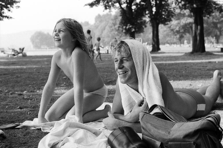 A imagem em preto e branco mostra uma criança e um adulto relaxando em um parque durante um dia ensolarado. A criança, com cabelo longo e solto, está de joelhos e sorrindo, enquanto o adulto, com cabelo loiro e enrolado, está deitado de lado, com uma toalha sobre os ombros e também sorrindo. Ao fundo, há outras pessoas e árvores, além de uma área de água visível.