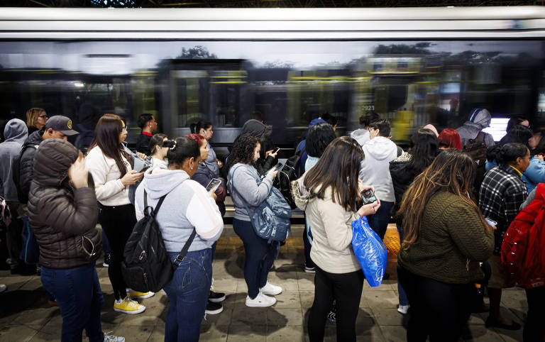 A imagem mostra um grupo de pessoas em uma estação de transporte público, aguardando a chegada de um veículo. A maioria das pessoas está olhando para seus celulares. Um ônibus urbano moderno passa rapidamente ao fundo, criando um efeito de movimento. As pessoas estão vestidas de forma casual, algumas com jaquetas e mochilas.