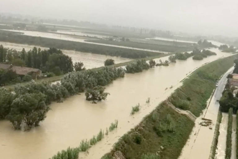 A imagem mostra uma vasta área rural inundada, com água cobrindo campos e árvores. O céu está nublado, indicando um clima chuvoso. À direita, há uma estrada que parece estar parcialmente submersa. A vegetação ao redor é densa, com árvores visíveis ao longo do curso da água.