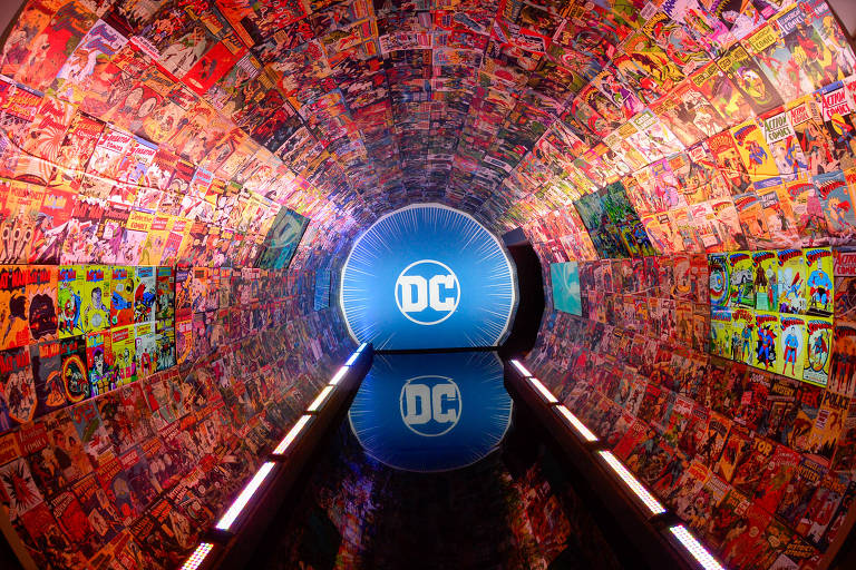 A imagem mostra um túnel decorado com capas de quadrinhos da DC Comics nas paredes. No fundo do túnel, há um círculo azul com o logotipo da DC. O chão reflete a luz, criando um efeito visual interessante.