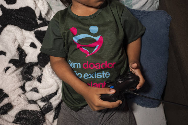 Uma criança está sentada segurando um controle de videogame. Ela usa uma camiseta verde com a frase 'Sem doador não existe transplante' em letras coloridas. Ao fundo, há uma manta com padrão de manchas pretas e brancas e um travesseiro azul.