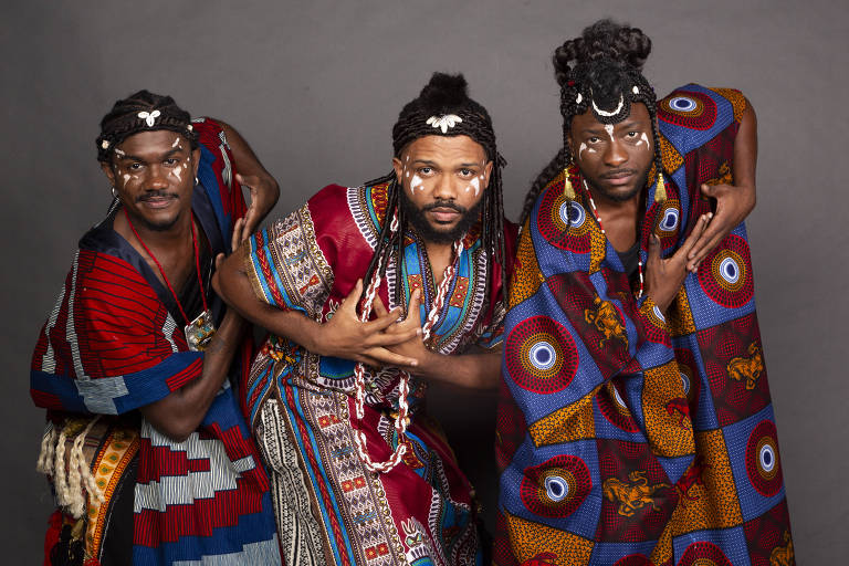 Três homens posam juntos, vestindo trajes tradicionais coloridos. O homem à esquerda usa uma roupa com listras vermelhas e azuis, adornado com colares. O homem central veste uma túnica vermelha com detalhes coloridos e tem pinturas brancas no rosto. O homem à direita usa um manto com padrões azuis e vermelhos, também com pinturas faciais. Todos têm cabelos trançados e expressões sérias, com um fundo cinza.