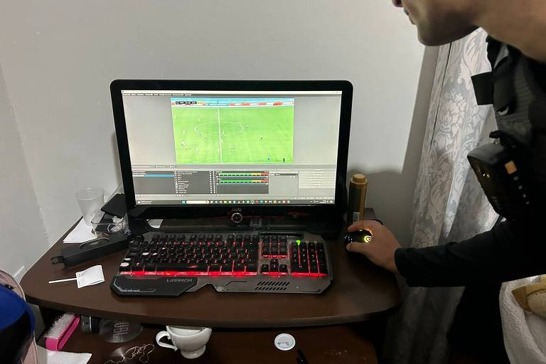 A imagem mostra um computador de mesa com um monitor exibindo um jogo de futebol. O teclado é iluminado em vermelho e há um mouse ao lado. Um homem, parcialmente visível, está ao lado do computador, usando uma camisa escura e um colete. Na mesa, há um copo, um recipiente branco, e outros objetos não identificáveis.