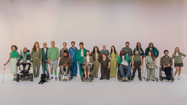 A imagem mostra um grupo diversificado de pessoas posando em um fundo neutro. O grupo é composto por homens e mulheres de diferentes idades e etnias, incluindo pessoas em cadeiras de rodas e com deficiência. Todos estão vestidos em tons de verde e apresentam uma variedade de estilos, desde roupas casuais até mais formais. O ambiente é iluminado de forma suave, destacando a diversidade e a inclusão.