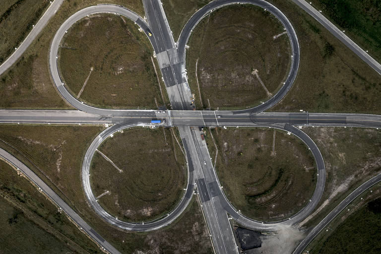 Imagem aérea de uma interseção rodoviária com quatro rotatórias dispostas em forma de flor. As rotatórias são circulares e conectadas por estradas retas. O terreno ao redor é predominantemente verde, com algumas áreas de asfalto visíveis