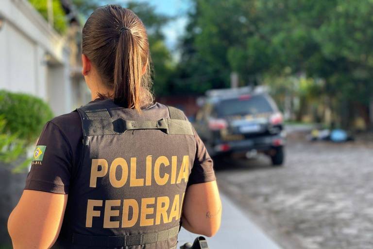 Uma policial de farda com um colete escrito POLÍCIA FEDERAL. Ela está de costas， o cabelo amarrado e no fundo uma viatura e uma casa.