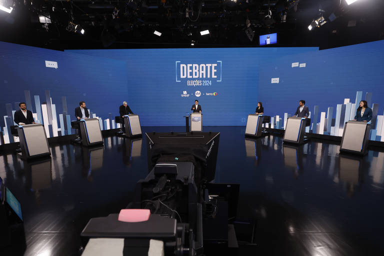 A imagem mostra um cenário de debate eleitoral com sete participantes em pé atrás de mesas. O fundo é azul com gráficos de barras brancas. No centro, uma mesa com um microfone está posicionada para o moderador. As luzes do estúdio estão acesas, criando um ambiente de discussão.