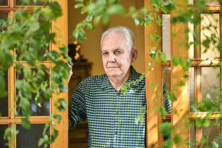 Um homem idoso está em pé, olhando para a câmera, enquanto se apoia em uma porta de madeira. Ele usa uma camisa xadrez verde e azul. Ao redor da porta, há folhagens verdes que emolduram a cena