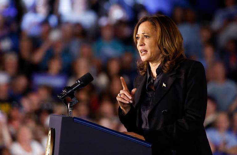 Foto mostra a vice-presidente dos EUA, Kamala Harris em um palanque falando ao microfone. Kamala é uma mulher negra de pele clara, cabelos lisos e castanhos. Ela veste um terno preto com camisa também preta e no seu blazer existe um broche dourado no formato da bandeira estadunidense. Kamala esta com o dedo indicador em riste e é possível ver um anel dourado nesta mão. Ao fundo, uma multidão assiste o discurso nas arquibancadas.