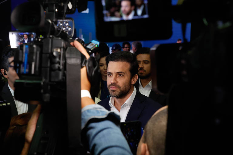 Homem de barba rala e cabelos curtos pretos, de camisa branca e paletó preto, está cercado por jornalistas e câmeras. Aparece no meio da foto, com uma grande câmera erguida no ar no canto direito da imagem. O cenário é sombrio