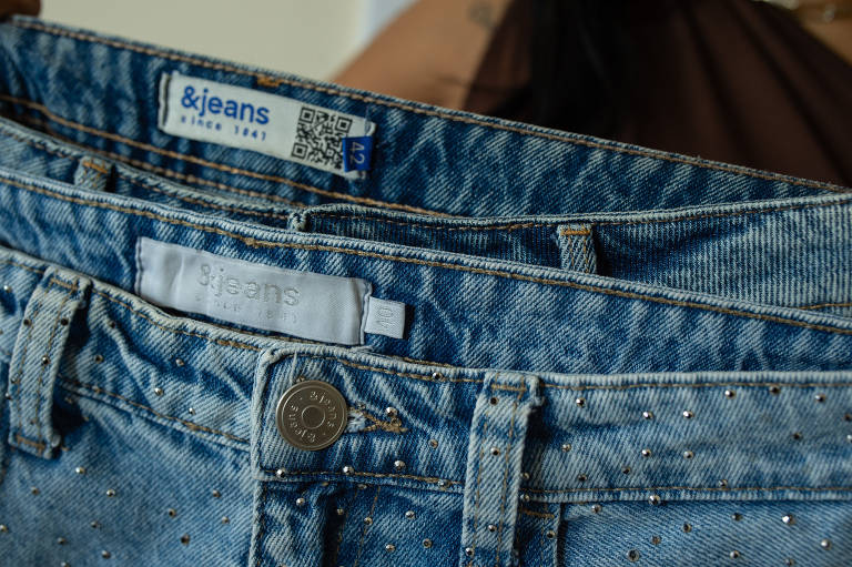 A imagem mostra uma calça jeans com detalhes em strass na parte frontal. A etiqueta visível indica o tamanho 40. No fundo há uma segunda peça, azul e cinza, de tamanho 42.