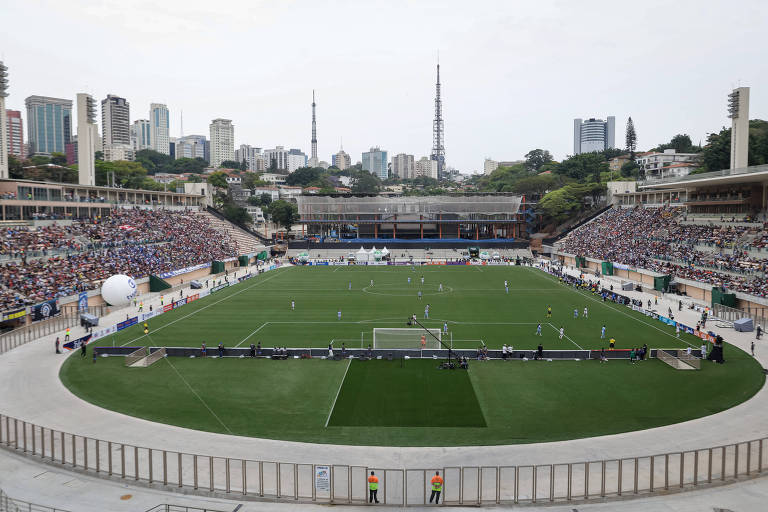 Imagem de um estádio de futebol com um campo verde e jogadores em ação. O público está assistindo nas arquibancadas， que estão cheias. Ao fundo， há prédios altos e torres de comunicação， indicando um ambiente urbano. O céu está nublado， e há uma estrutura em construção visível.
