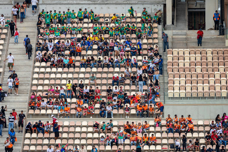 A imagem mostra um estádio com várias arquibancadas. Há torcedores sentados em diferentes seções, com destaque para grupos usando camisetas de cores variadas, como verde, amarelo, laranja e vermelho. Algumas áreas estão mais cheias, enquanto outras estão quase vazias. O ambiente parece ser de um evento esportivo, com pessoas se movimentando nas escadas e nas arquibancadas.