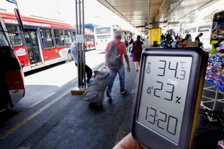 A imagem mostra um termômetro digital em primeiro plano marcando temperatura de 34，3ºC e umidade de 23% às 13h20 no terminal Parque Dom Pedro 