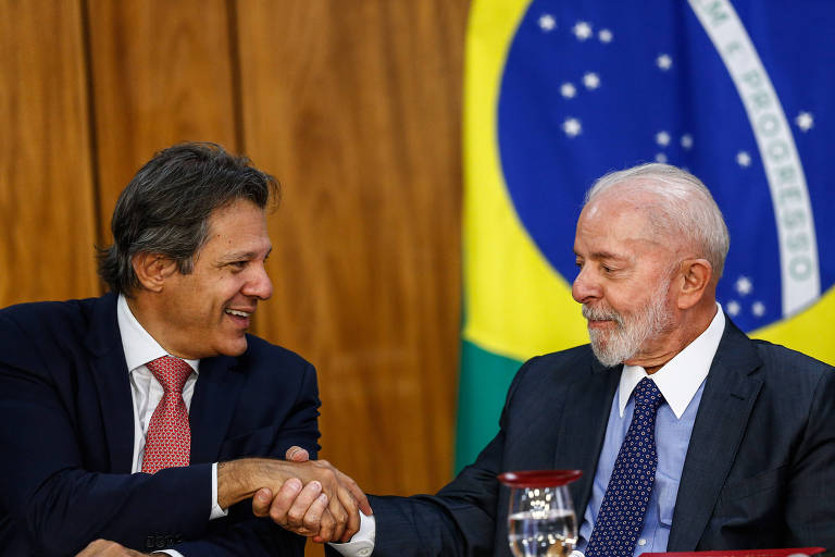 Dois homens estão se cumprimentando com um aperto de mão. O homem à esquerda está sorrindo e usa um terno escuro, enquanto o homem à direita, também sorrindo, veste um terno claro e uma gravata escura. Ao fundo, há uma bandeira do Brasil visível, com as cores verde, amarelo e azul.
