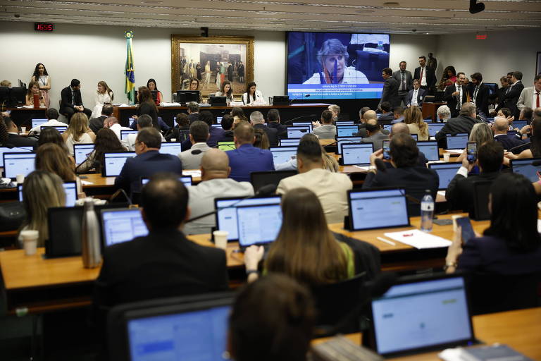 A imagem mostra uma sala de reuniões com várias pessoas sentadas em mesas， cada uma utilizando laptops. No fundo， há uma tela grande exibindo uma pessoa falando. A sala é iluminada e decorada com uma pintura ao fundo， além de uma bandeira do Brasil visível. O ambiente parece ser formal e está relacionado a uma sessão legislativa.
