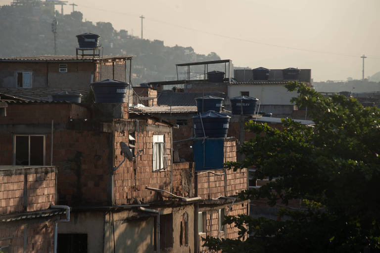 A imagem mostra uma vista de várias edificações em uma área urbana， com casas de tijolos expostos e tanques de água azuis no telhado. A iluminação é suave， sugerindo um horário próximo ao amanhecer ou entardecer. Ao fundo， há uma colina e algumas estruturas industriais ou de telecomunicações visíveis.