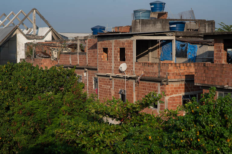 A imagem mostra uma construção de tijolos expostos, com janelas sem vidro e algumas paredes inacabadas. No telhado, há tanques azuis e uma antena parabólica visível. A vegetação densa na parte inferior da imagem contrasta com as estruturas urbanas ao fundo. O céu está claro, indicando um dia ensolarado.