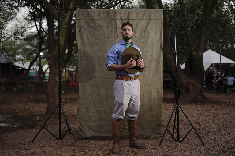 Um homem está posando em frente a um fundo de tecido verde em um ambiente ao ar livre. Ele usa uma camisa azul, um lenço verde no pescoço, calças claras e botas altas. O cenário é cercado por árvores e há tendas ao fundo, sugerindo um evento ao ar livre.