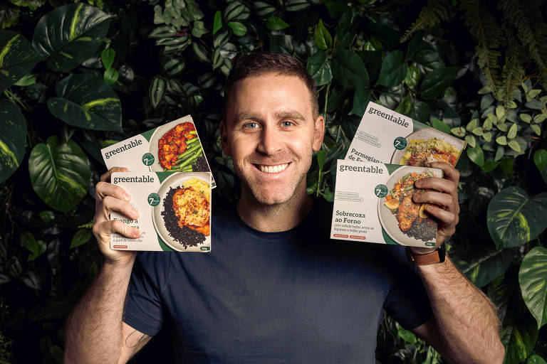 Um homem sorridente está posando em frente a um fundo de folhas verdes. Ele segura quatro embalagens de comida， duas em cada mão， mostrando diferentes pratos. O homem está vestindo uma camiseta escura e tem cabelo curto.